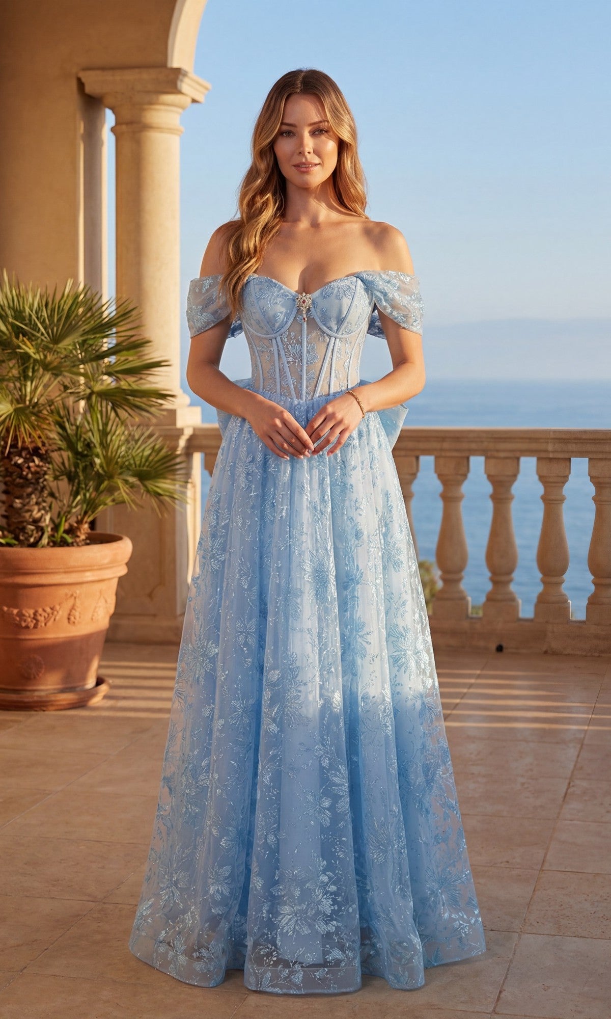 A woman stands on a terrace with the ocean and palms behind her, wearing the blue Long Prom Dress: Chicas C391—an off-the-shoulder, floor-length gown with a sheer corset bodice and floral overlay.